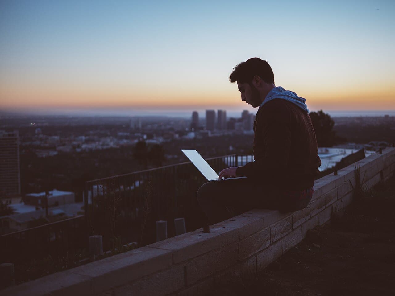 A traveler working on a laptop from a city viewpoint at sunset.